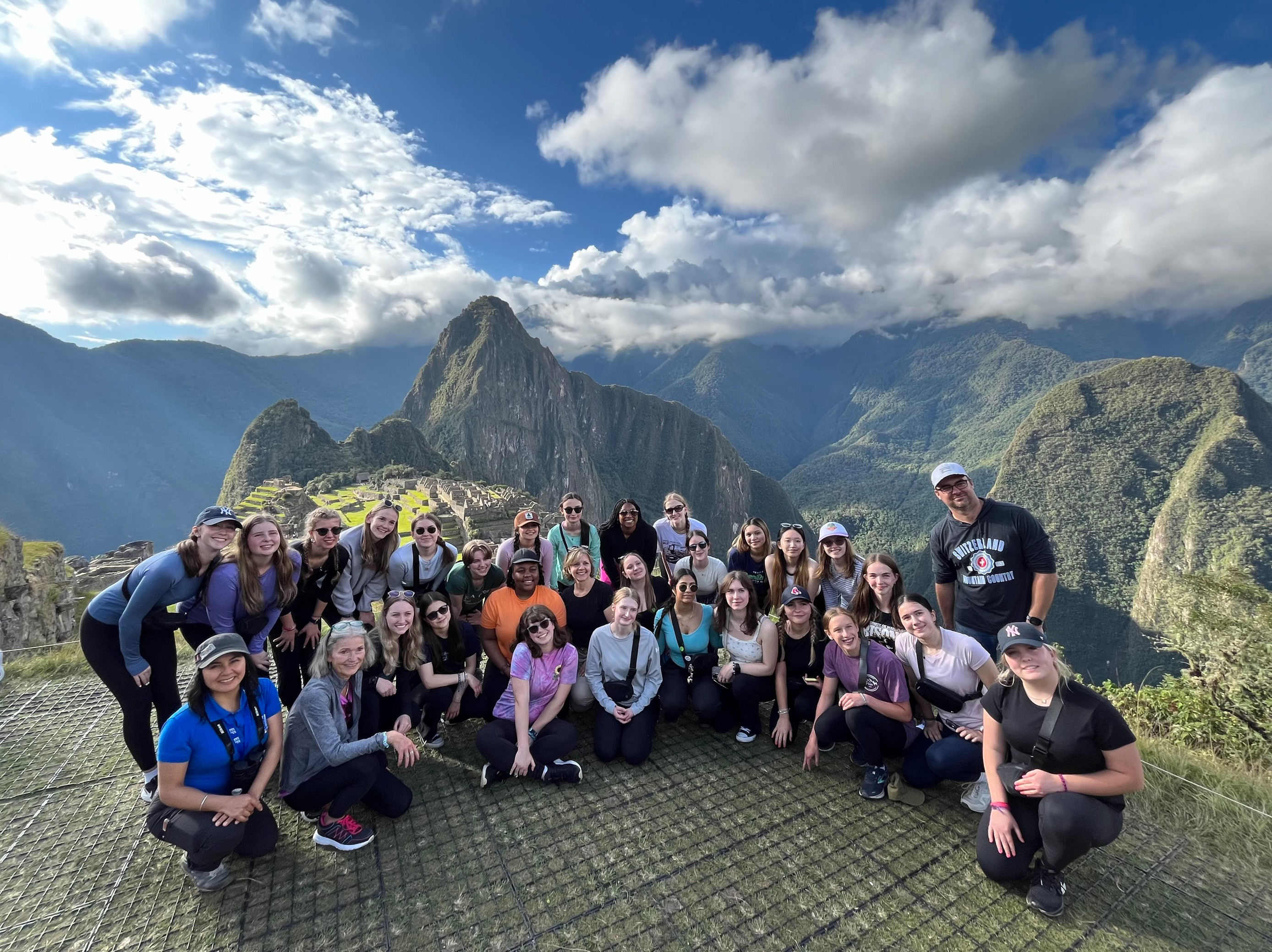 A group of smiling students and adult chaperones stand in front of a mountain with wisps of clouds encircling it.